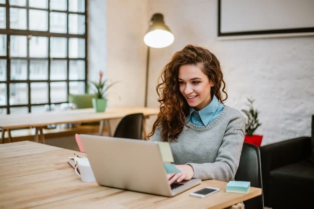 Una mujer con cabello rizado está trabajando en una computadora portátil en un escritorio de madera en una habitación luminosa con una ventana grande y plantas de interior, posiblemente participando en alguna actividad de networking.