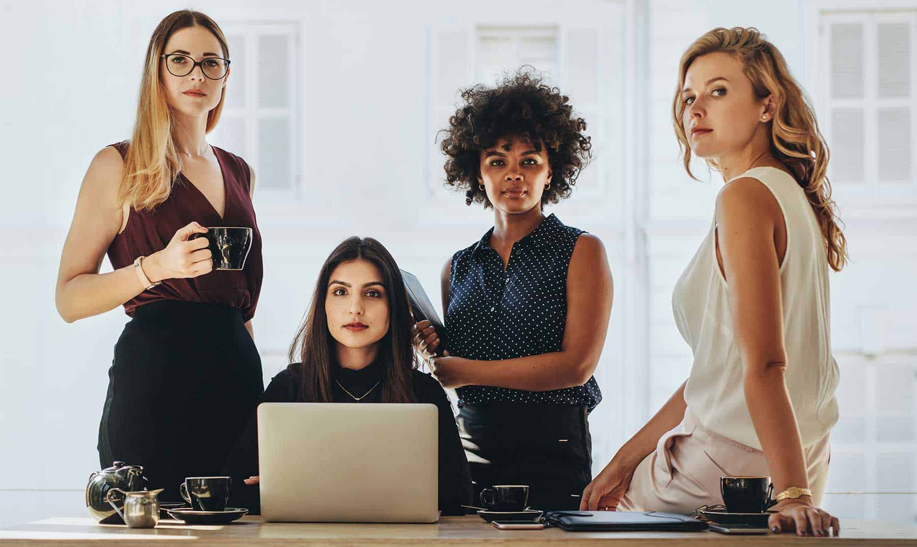 Cuatro mujeres están reunidas alrededor de una mesa, una sentada con un ordenador portátil y tres de pie. Las mujeres parecen estar en un entorno profesional, probablemente participando en actividades de networking.