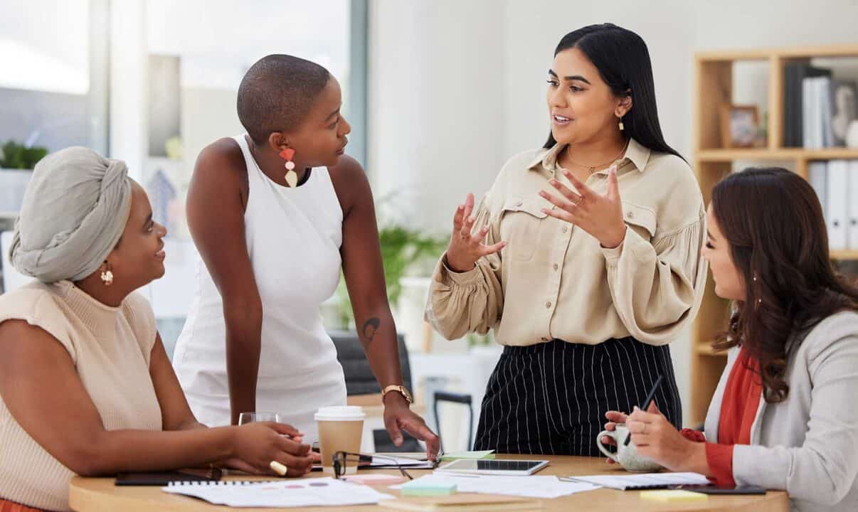 Un grupo de cuatro mujeres mantiene una conversación en una oficina. Una mujer está de pie y gesticula con las manos mientras las demás están sentadas y escuchan atentamente. Sobre la mesa hay varios documentos y una taza de café, lo que ilustra una sesión de networking productiva.