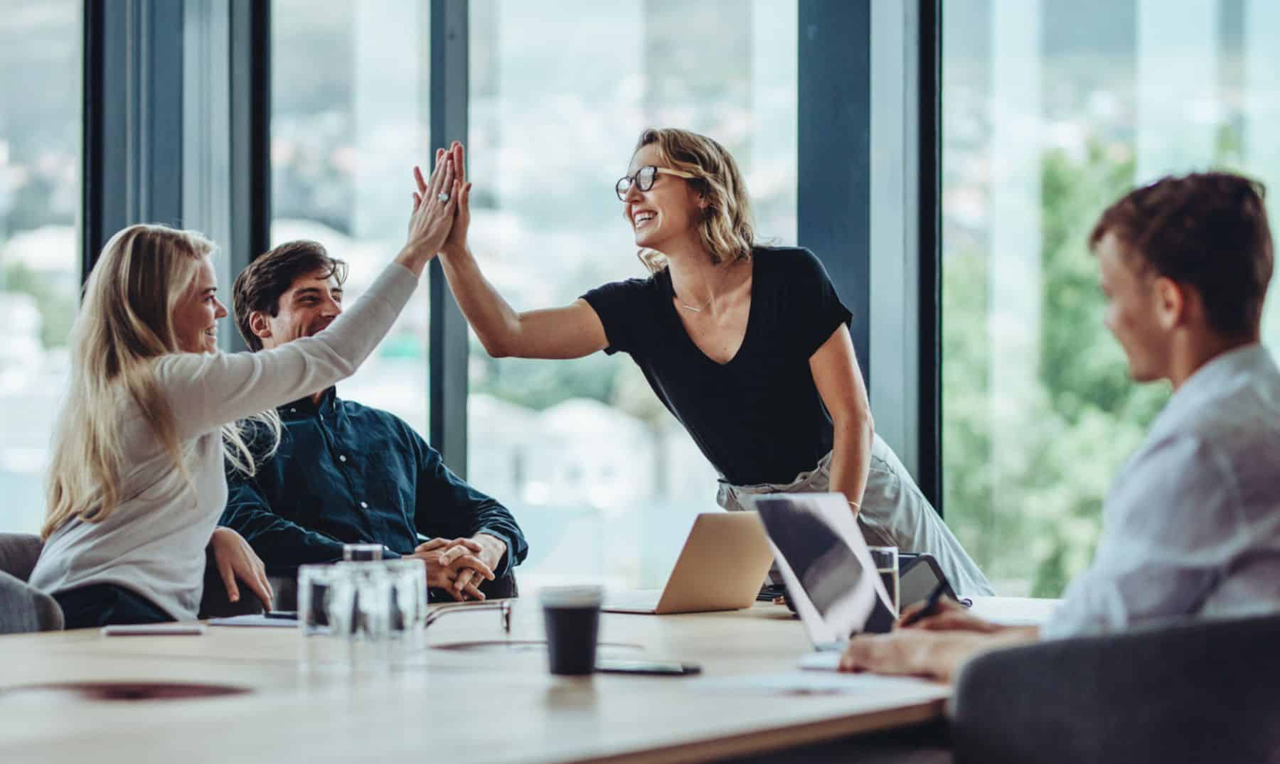 En una oficina moderna, cuatro colegas se encuentran haciendo networking y dos de ellos chocan los cinco para celebrar un momento exitoso, mientras los demás observan y continúan trabajando en sus computadoras portátiles.