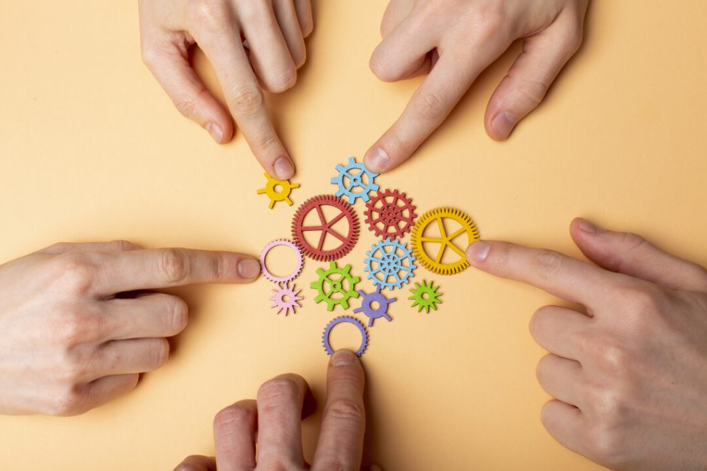 Cuatro manos apuntan a engranajes de colores interconectados sobre una superficie beige, que simbolizan el trabajo en equipo, la colaboración y la adaptación y manejo efectivo de cambios.