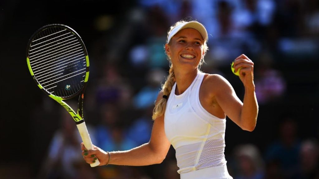 Una sonriente Caroline Wozniacki, vestida con un traje blanco, sostiene su raqueta y una pelota de tenis en una cancha al aire libre soleada con espectadores al fondo.
