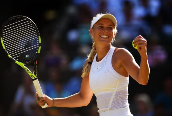 Una sonriente Caroline Wozniacki, vestida con un traje blanco, sostiene su raqueta y una pelota de tenis en una cancha al aire libre soleada con espectadores al fondo.