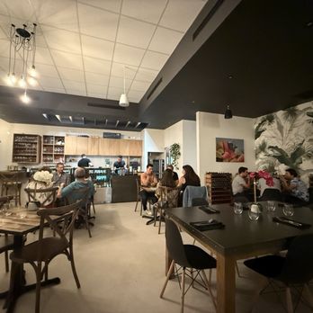 El interior de un moderno restaurante en Edan Bistro presenta varios grupos de personas cenando, mesas y sillas de madera y una vista de la bulliciosa cocina abierta al fondo.