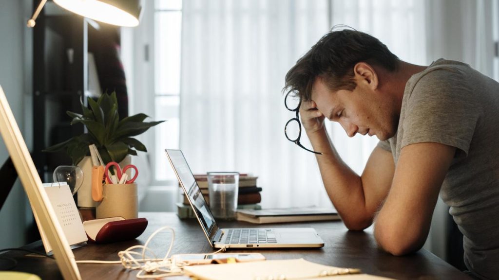 Un hombre se sienta en un escritorio con la cabeza entre las manos, con aspecto estresado mientras trabaja en una laptop. Rodeado de material de oficina, una planta y un vaso de agua, piensa en maneras de evitar el agotamiento emocional.