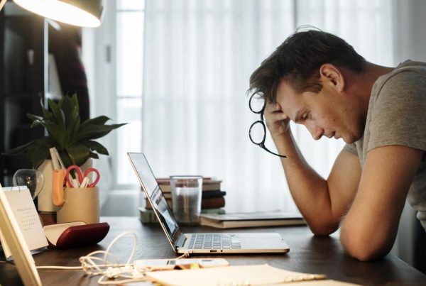 Un hombre se sienta en un escritorio con la cabeza entre las manos, con aspecto estresado mientras trabaja en una laptop. Rodeado de material de oficina, una planta y un vaso de agua, piensa en maneras de evitar el agotamiento emocional.