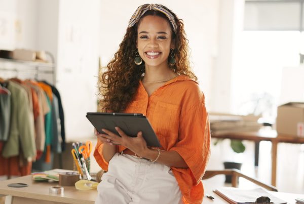 Una mujer con una blusa naranja está de pie en un espacio de trabajo, sonriendo y sosteniendo una tableta mientras se prepara para empezar. Al fondo se ven ropa y material de oficina.
