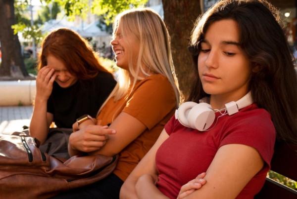 Tres chicas adolescentes se sientan en un banco; mientras dos se ríen juntas, la tercera, con auriculares, se sienta aparte con los brazos cruzados y parece molesta, una imagen que insinúa posibles amistades tóxicas.