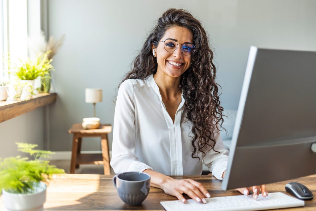 Una mujer con cabello largo y rizado y gafas se sienta en un escritorio, sonriendo a un monitor de computadora, lista para incorporar hábitos que impulsan el éxito, con una taza de café y plantas cerca en un espacio de trabajo moderno y luminoso.