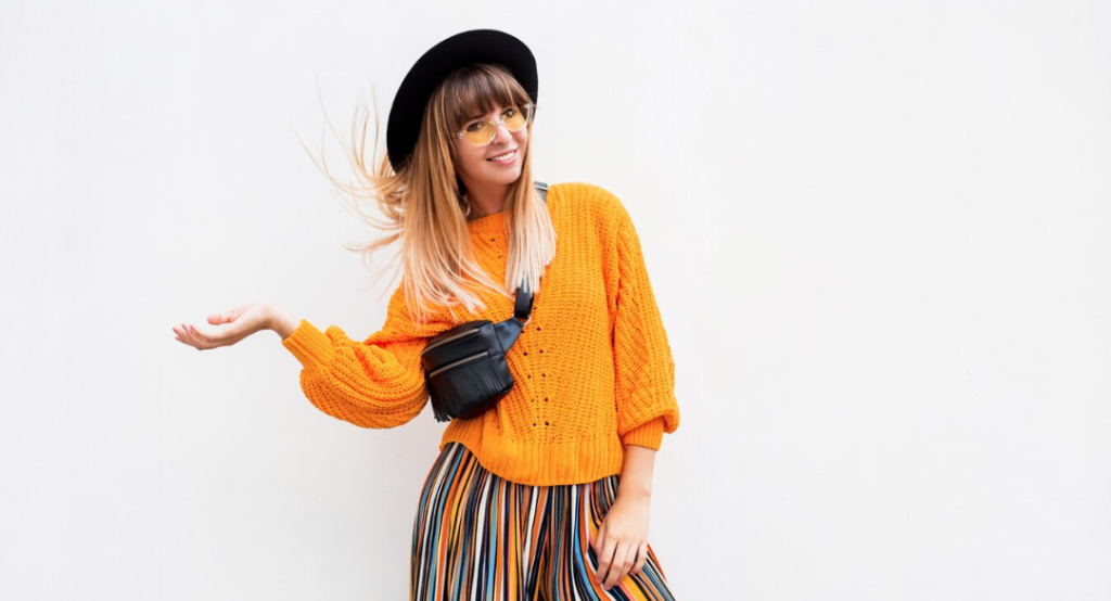 Una mujer que viste un suéter naranja, un sombrero negro, gafas amarillas, pantalones a rayas y una bolsa cruzada negra se encuentra sobre un fondo blanco liso, mostrando las tendencias de moda para otoño-invierno 2024 con confianza y estilo.