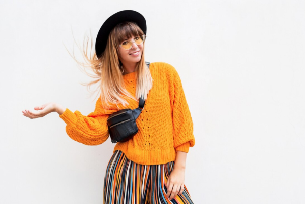 Una mujer que viste un suéter naranja, un sombrero negro, gafas amarillas, pantalones a rayas y una bolsa cruzada negra se encuentra sobre un fondo blanco liso, mostrando las tendencias de moda para otoño-invierno 2024 con confianza y estilo.