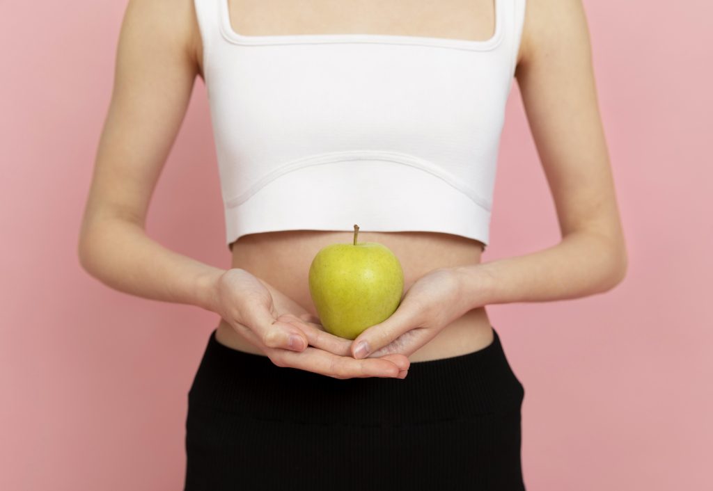 Una persona que viste un top blanco y pantalones negros sostiene una manzana verde (uno de los alimentos para mejorar la digestión) con ambas manos sobre un fondo rosa.