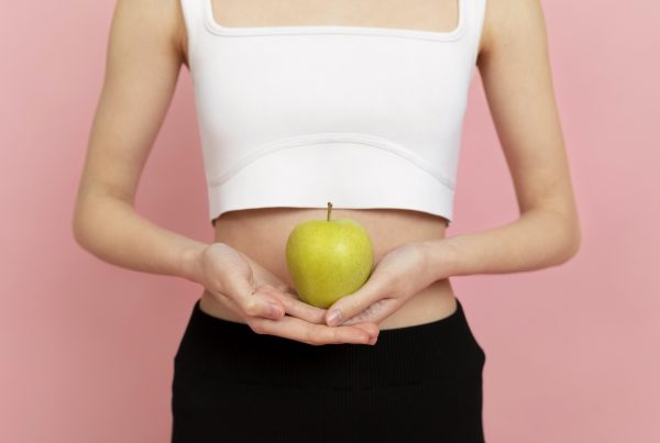 Una persona que viste un top blanco y pantalones negros sostiene una manzana verde (uno de los alimentos para mejorar la digestión) con ambas manos sobre un fondo rosa.
