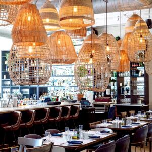 Un interior de restaurante moderno en Doya, la mejor comida del Egeo en Miami, con lámparas colgantes de mimbre, una barra llena de botellas y mesas preparadas con platos, vasos y utensilios.