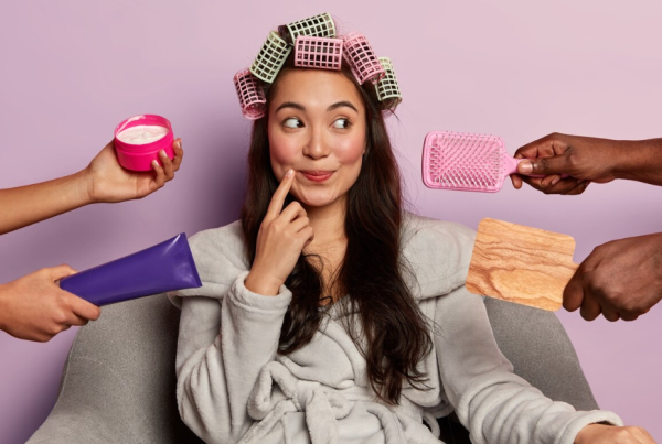 Una mujer con rulos se sienta en una silla mientras tres manos le ofrecen un cepillo para el cabello, crema facial y una paleta de madera sobre un fondo rosa, resaltando los mitos y realidades sobre la salud femenina.
