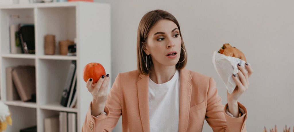 Una mujer con un blazer color melocotón está sentada en un escritorio, sosteniendo una manzana y un sándwich envuelto, sopesando cuidadosamente sus opciones, ilustrando el clásico dilema entre el hambre física y el hambre emocional.