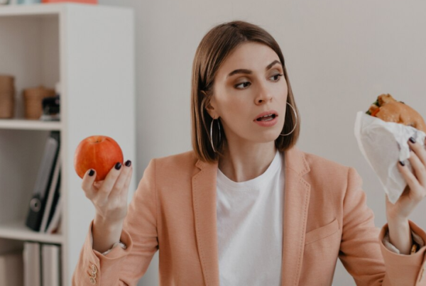 Una mujer con un blazer color melocotón está sentada en un escritorio, sosteniendo una manzana y un sándwich envuelto, sopesando cuidadosamente sus opciones, ilustrando el clásico dilema entre el hambre física y el hambre emocional.