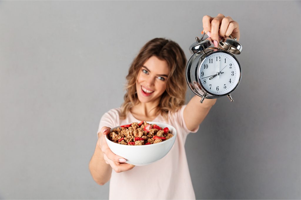 Mujer sonriendo, sosteniendo un tazón de granola con fruta en una mano y un reloj despertador analógico en la otra, de pie sobre un fondo gris liso, que representa el ayuno intermitente.