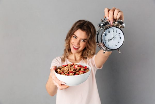 Mujer sonriendo, sosteniendo un tazón de granola con fruta en una mano y un reloj despertador analógico en la otra, de pie sobre un fondo gris liso, que representa el ayuno intermitente.