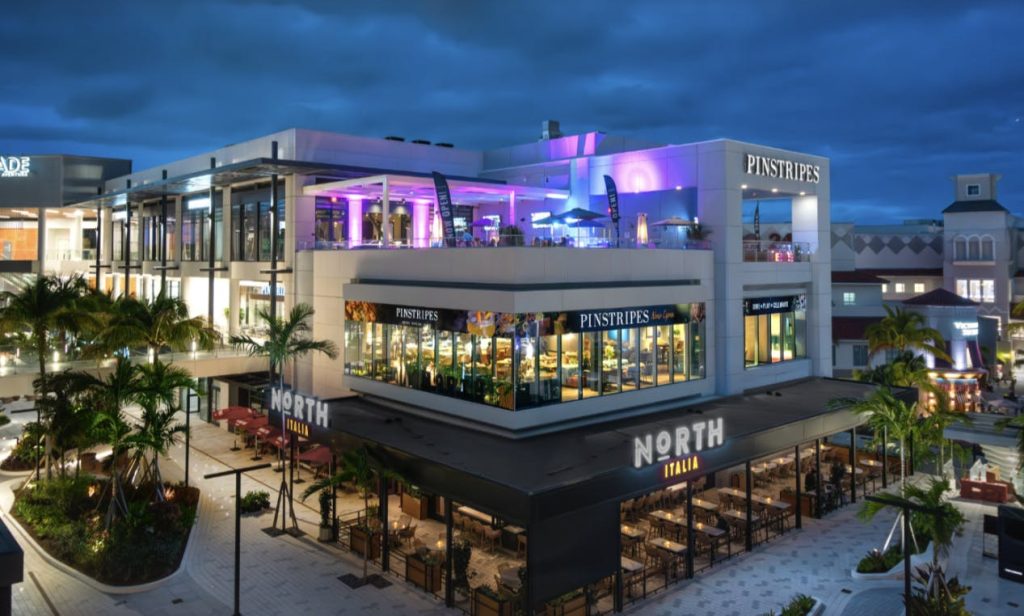 Un moderno edificio de varios niveles cuenta por la noche con los restaurantes Pinstripes Aventura y North Italia iluminados, con palmeras y asientos al aire libre visibles.