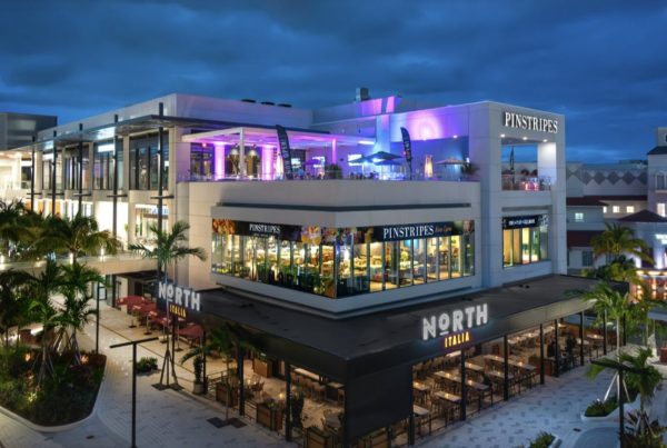 Un moderno edificio de varios niveles cuenta por la noche con los restaurantes Pinstripes Aventura y North Italia iluminados, con palmeras y asientos al aire libre visibles.