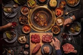 Una comida de barbacoa coreana en Cote Steakhouse incluye carnes crudas, guarniciones y salsas dispuestas alrededor de una parrilla, mientras la gente cocina y disfruta del festín juntos en la mesa.