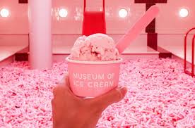 Una mano sostiene un vaso de helado con una cuchara, con la etiqueta "El Museo del Helado", en un ambiente bien iluminado y de temática rosa.