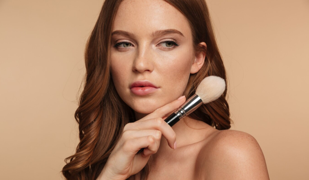 Una mujer con cabello largo castaño rojizo sostiene un pincel de maquillaje en su rostro, mirando ligeramente hacia un lado contra un fondo beige, mostrando maquillaje para un look natural y fresco.