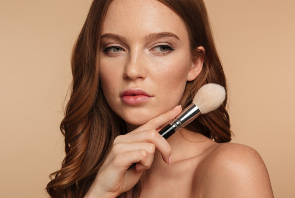 Una mujer con cabello largo castaño rojizo sostiene un pincel de maquillaje en su rostro, mirando ligeramente hacia un lado contra un fondo beige, mostrando maquillaje para un look natural y fresco.