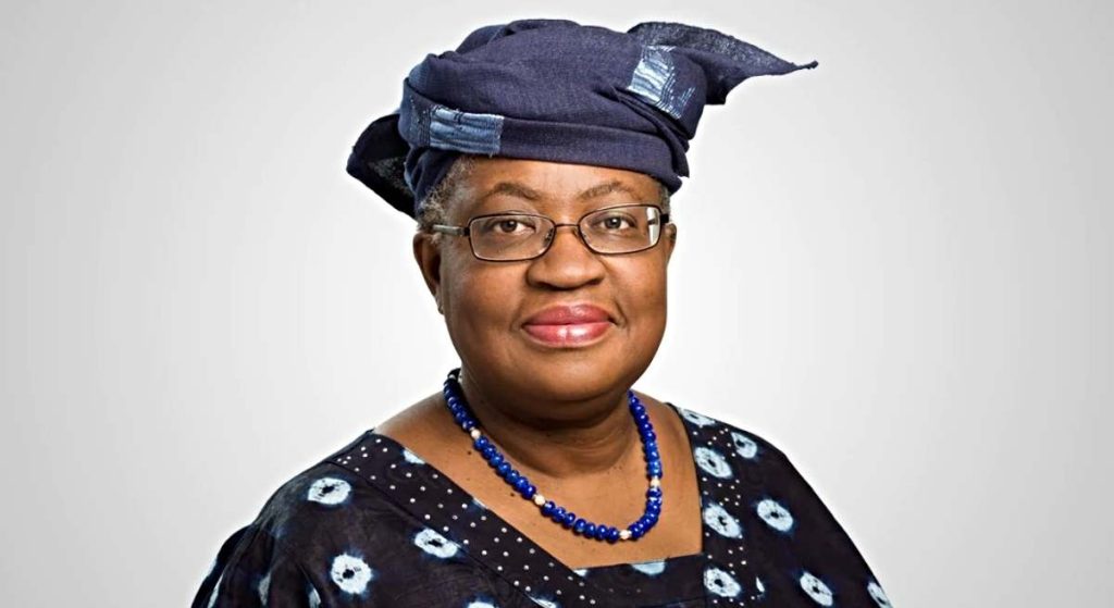 Una mujer que recuerda a Ngozi Okonjo-Iweala lleva gafas, un pañuelo azul oscuro estampado, una blusa a juego y un collar de cuentas azules mientras posa sobre un fondo claro y liso.