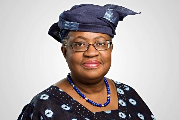 Una mujer que recuerda a Ngozi Okonjo-Iweala lleva gafas, un pañuelo azul oscuro estampado, una blusa a juego y un collar de cuentas azules mientras posa sobre un fondo claro y liso.