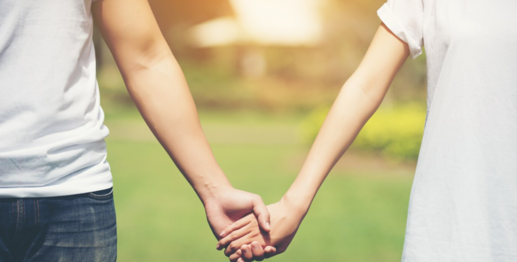 Dos personas paradas al aire libre se toman de la mano, resaltando sus manos y brazos unidos contra un fondo verde borroso, un gesto simple para fortalecer la relación con la pareja.