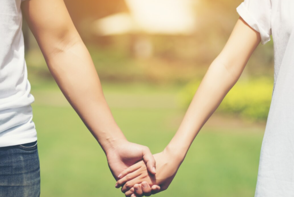 Dos personas paradas al aire libre se toman de la mano, resaltando sus manos y brazos unidos contra un fondo verde borroso, un gesto simple para fortalecer la relación con la pareja.