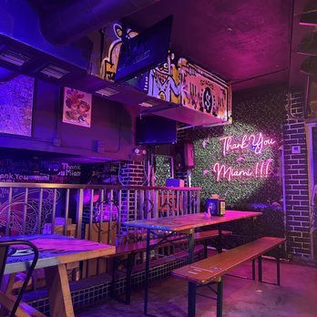 Bar interior en Thank You Miami Kitchen & Beerhouse con mesas y bancos de madera, letrero de neón "Thank You Miami!!" en una pared verde, murales coloridos e iluminación violeta vibrante.