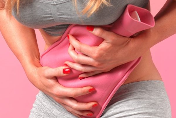 Una persona vestida de gris sostiene una bolsa de agua caliente de color rosa contra su abdomen inferior, lo que sugiere alivio del dolor de estómago o menstrual, posiblemente debido a la endometriosis.
