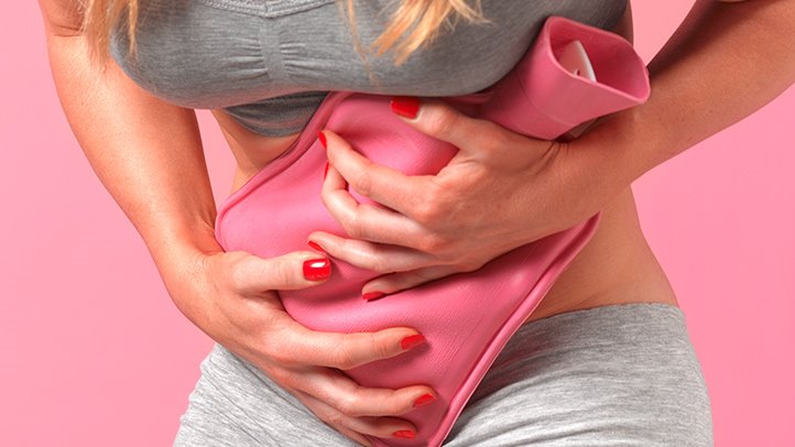 Una persona vestida de gris sostiene una bolsa de agua caliente de color rosa contra su abdomen inferior, lo que sugiere alivio del dolor de estómago o menstrual, posiblemente debido a la endometriosis.