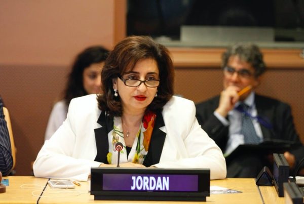 Una mujer vestida formal, parecida a Sima Bahous, está sentada en una mesa de conferencias con un micrófono, una placa que dice "JORDAN" y gente sentada en el fondo.