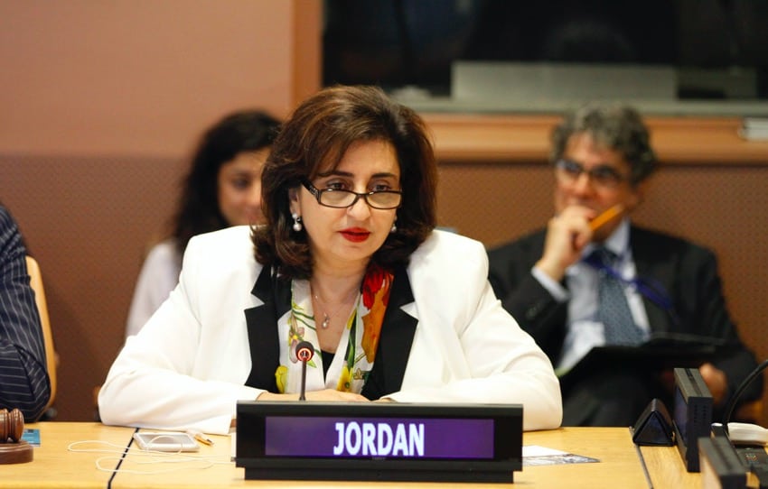 Una mujer vestida formal, parecida a Sima Bahous, está sentada en una mesa de conferencias con un micrófono, una placa que dice "JORDAN" y gente sentada en el fondo.