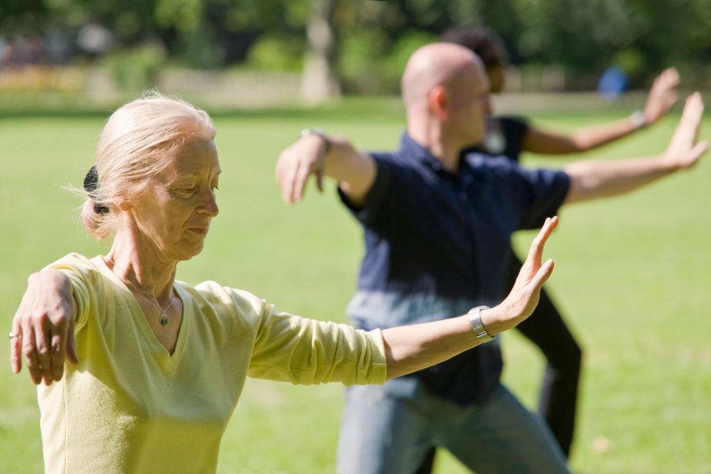 Tres adultos se encuentran en un parque con césped practicando tai chi, con los brazos levantados y extendidos frente a ellos, demostrando flexibilidad con la edad.